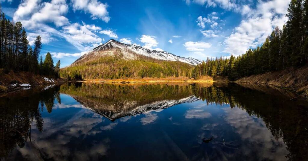 Panoramic view of a snow-capped mountain reflected in a clear, still lake surrounded by a forest under a partly cloudy blue sky.