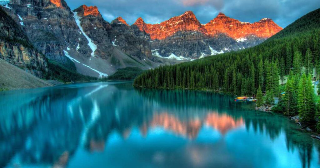 Early morning view of a serene lake reflecting the vivid, fiery red peaks of a rugged mountain range, surrounded by lush green forests under a clear blue sky.