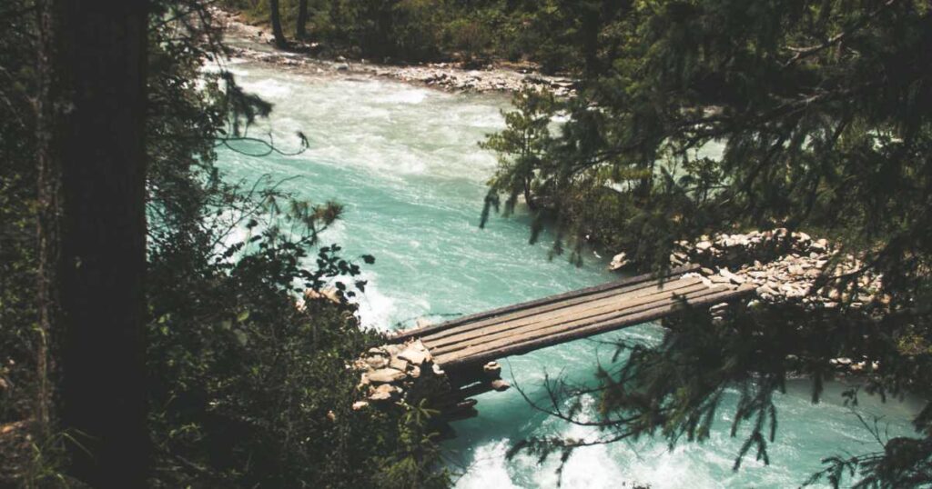 A narrow wooden bridge crosses a turquoise river, surrounded by dense forest.