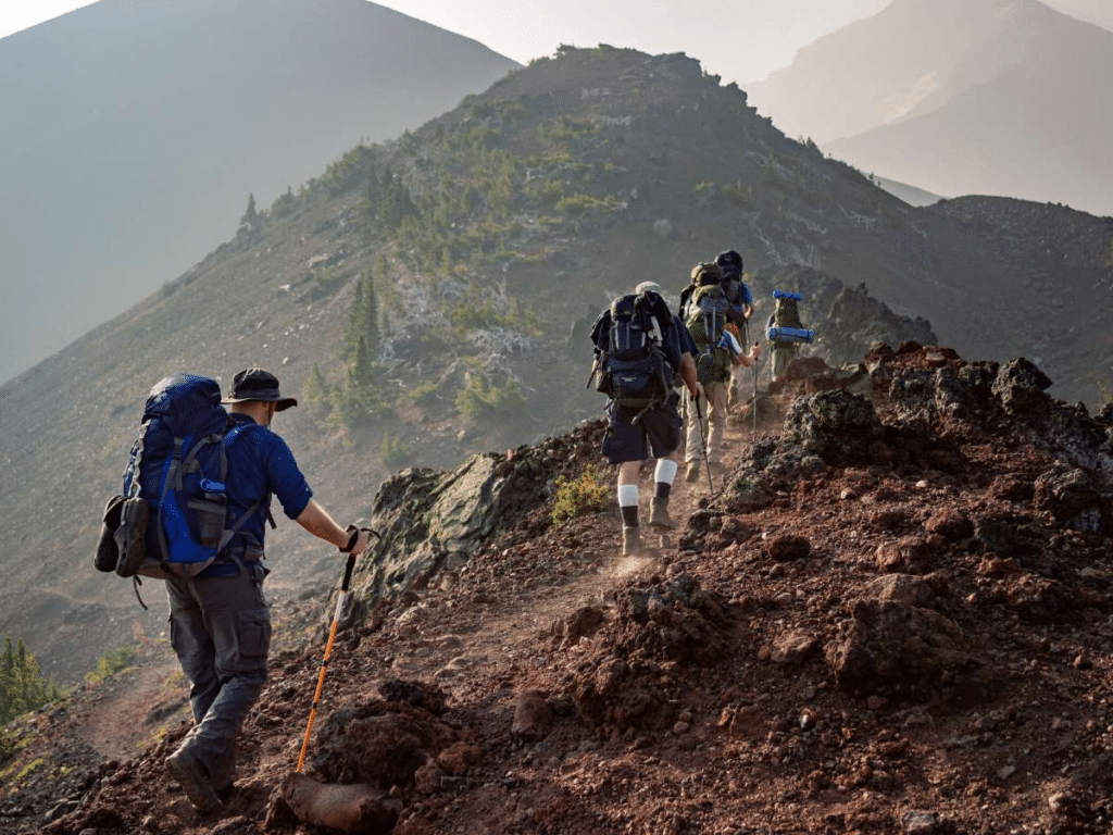 A group of hikers with backpacks trekking up a rugged mountain trail, surrounded by a mix of barren and vegetated slopes, under a hazy sky that suggests early morning light or dust in the air.
