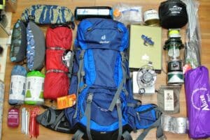 A variety of hiking essentials neatly laid out next to a blue Deuter backpack, including a sleeping bag, water bottles, camera, journal, and camping gear, ready for an outdoor adventure