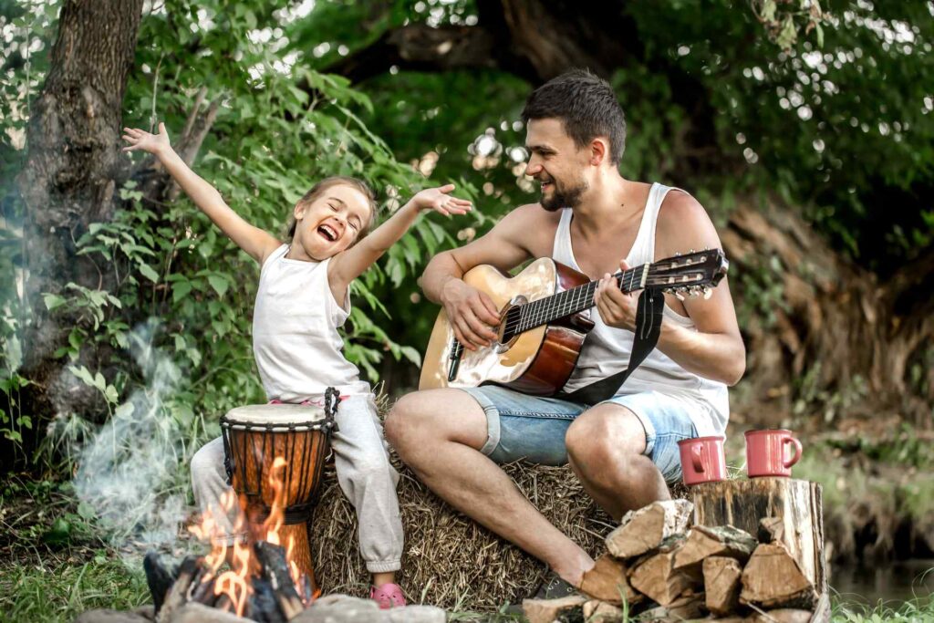 A joyful moment captured as a father and his young daughter enjoy music by a campfire outdoors. The father, wearing a tank top, plays a guitar, while his daughter, in a white sleeveless top, excitedly throws her arms in the air, sitting beside a drum
