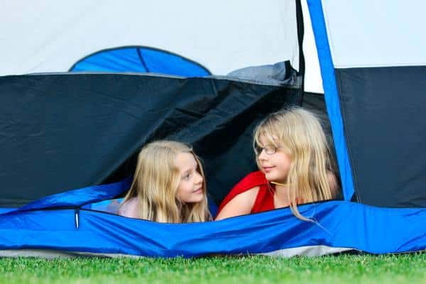 Two young girls smiling at each other inside a blue camping tent, with the tent flap open, set on a grassy field
