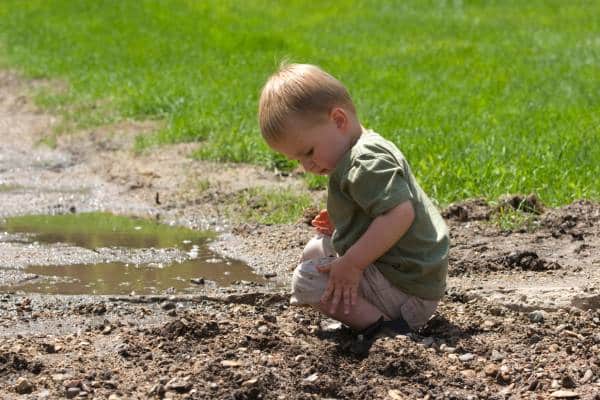 A toddler in a green shirt kneels next to a muddy puddle, playing with mud on a sunny day in a grassy area.