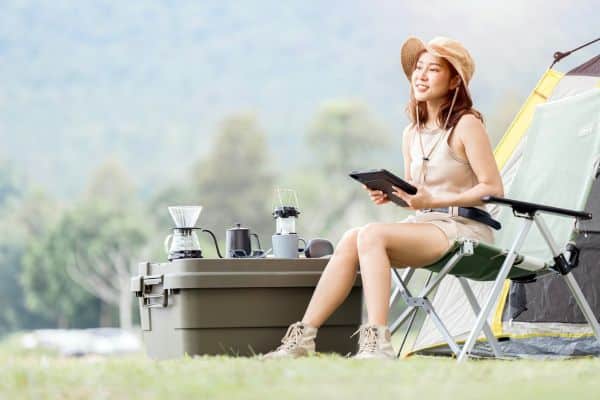 A woman in a sleeveless top and hat sits contentedly in a camping chair, reading a tablet with a coffee setup on a cooler next to her, signifying solo camping in a lush green environment