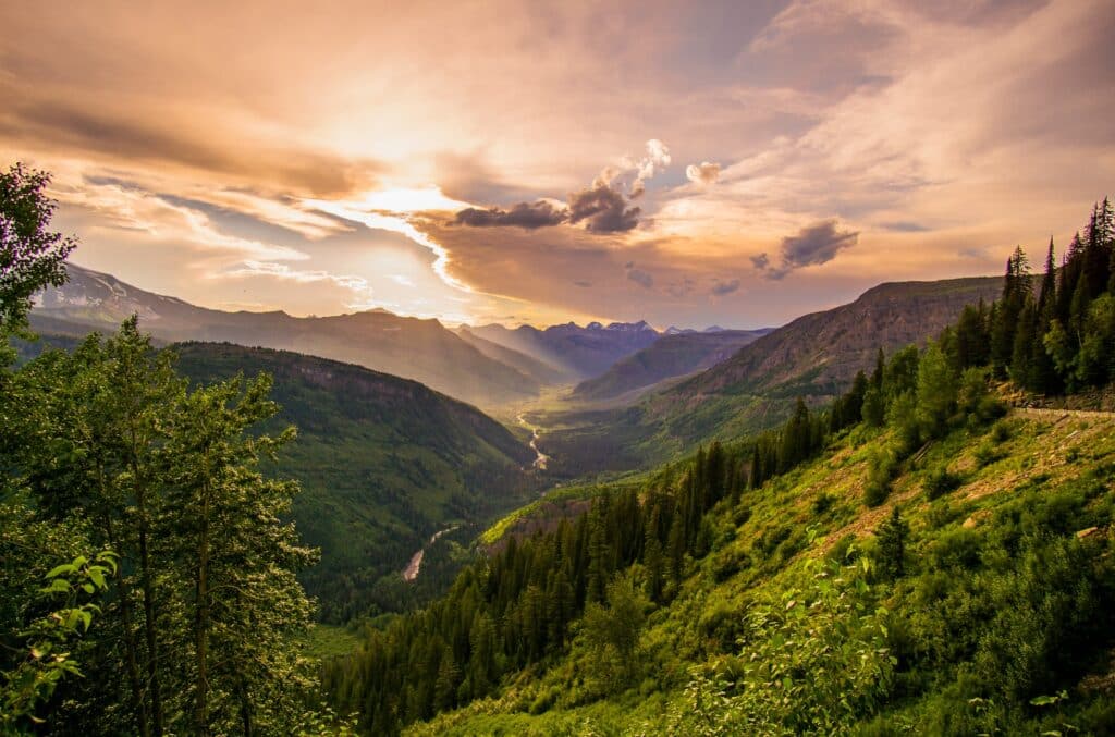 Sunset casting a golden glow over a lush mountain valley with dense forests and dynamic cloud formations.