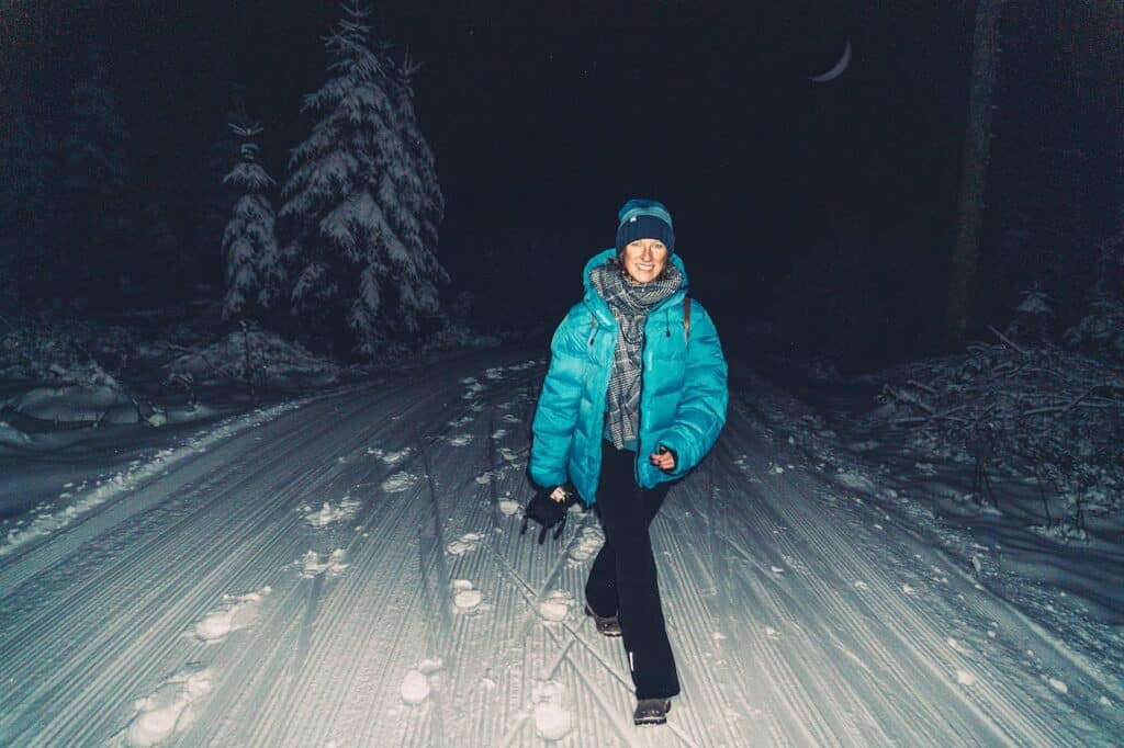 A person in a blue winter jacket and beanie smiling at the camera during a night-time walk on a snow-covered path, with the moon partially visible through the trees in the background