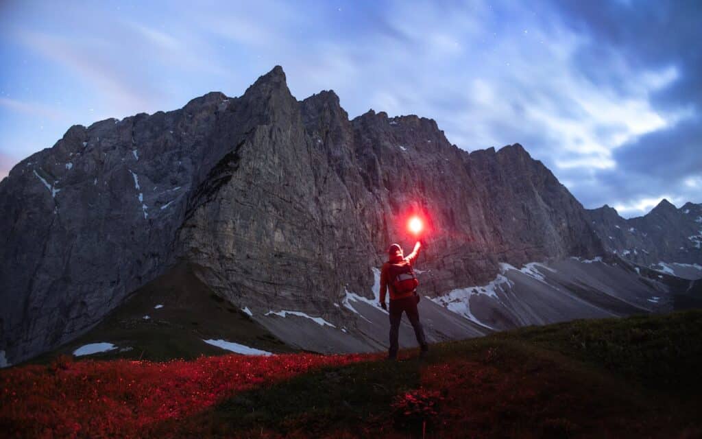 An adventurer holding up a red flare against the twilight sky on a mountainous terrain, with the imposing face of a rocky mountain in the background and a field of red flowers in the foreground.