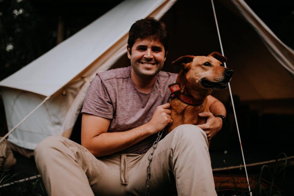 A cheerful man with a slight beard and wearing a maroon t-shirt cuddles with his happy brown dog wearing a red collar. They are sitting in front of a canvas tent, implying a joyful camping experience in a natural setting