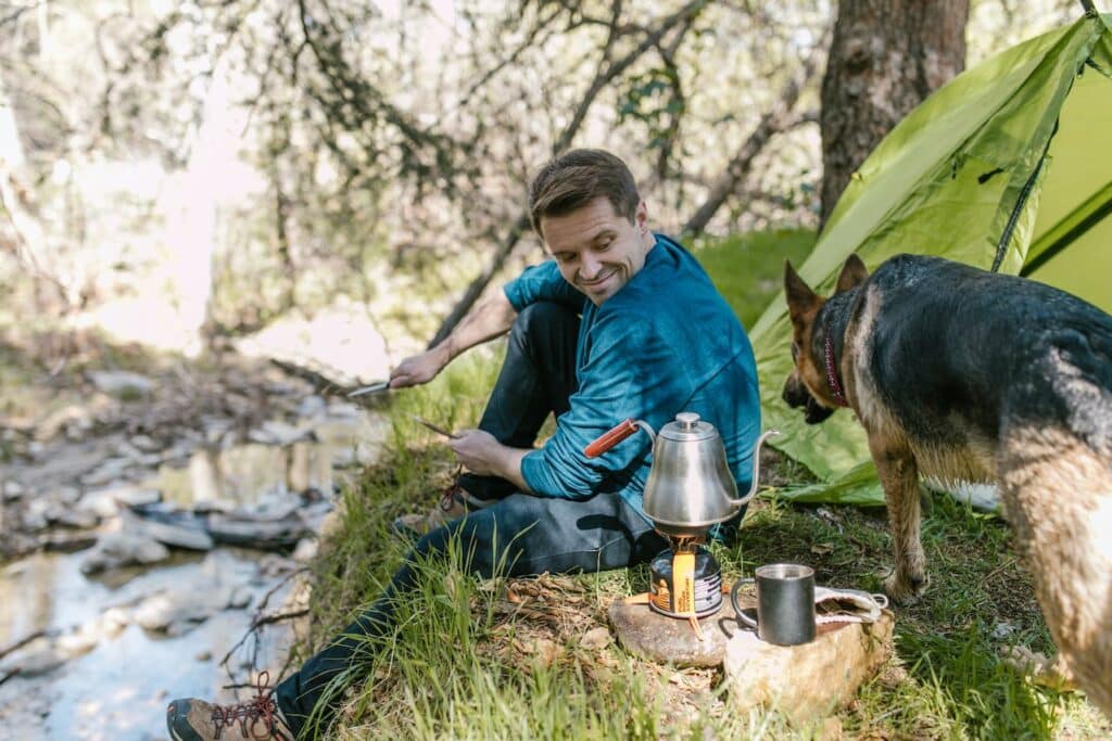 A man in a blue shirt smiles while seated on the ground by a green tent, preparing food on a portable stove with a kettle atop it. A German Shepherd dog stands beside him, attentively looking away, possibly at the serene woodland and stream in the background. The scene exudes a sense of peaceful outdoor camping