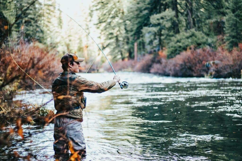 An angler in camouflage attire casts a fishing line into a serene river, surrounded by lush forest, capturing the tranquility and focus of fly fishing.