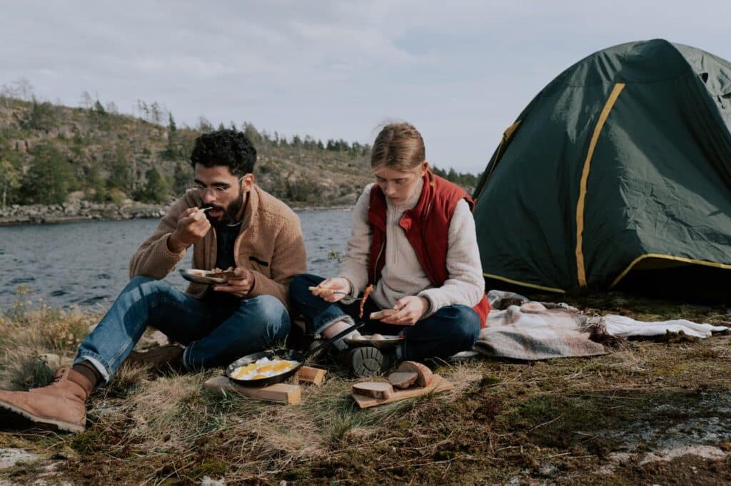 Two people enjoying a meal by a lake with a green and yellow tent in the background. The setting suggests a serene camping scene with one person eating from a bowl and the other spreading jam on bread, both seated on the ground.