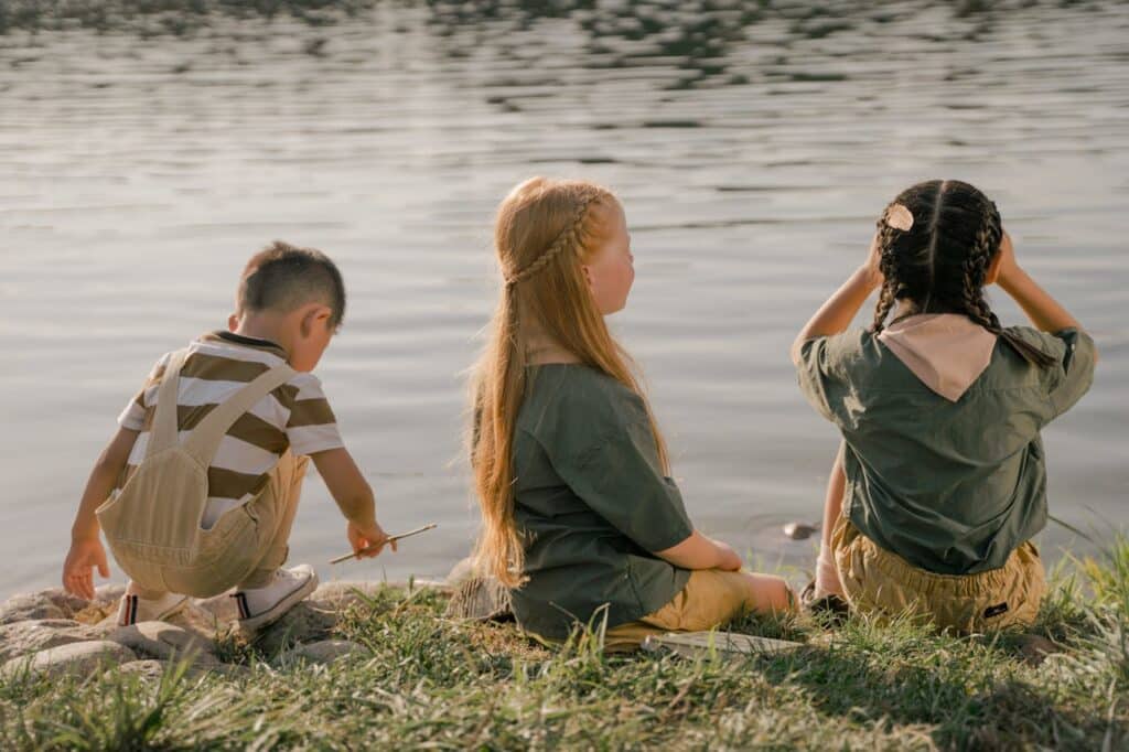 Three children sitting by a calm lake, with the focus on a girl with braided hair and a boy playing with a stick, while another girl looks on. They appear engaged in a peaceful lakeside activity, enjoying the outdoors.