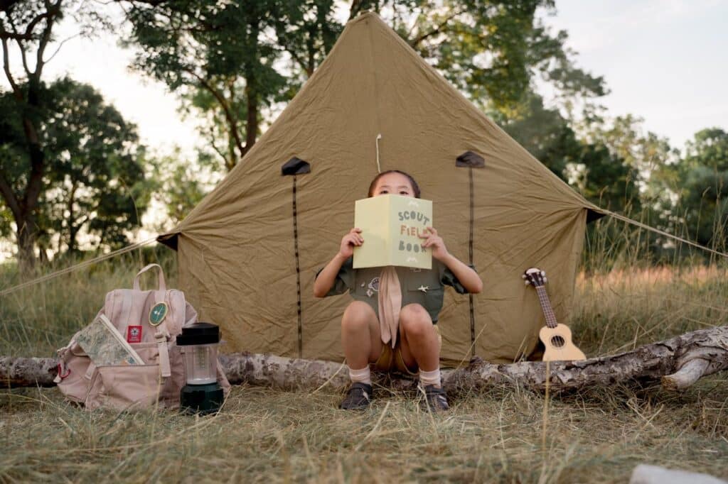 A child in a scout uniform sits in front of a beige tent, holding up a 'Scout Field Book' in front of their face. A backpack with badges, a lantern, and a ukulele lie nearby, contributing to a classic camping and scouting ambiance