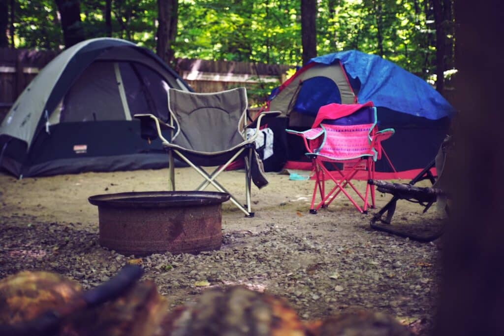 A campsite scene featuring two tents, one grey and one blue with a tarp, and a variety of camping chairs around a metal fire ring. The presence of camping gear and the surrounding dense trees suggest a peaceful outdoor retreat