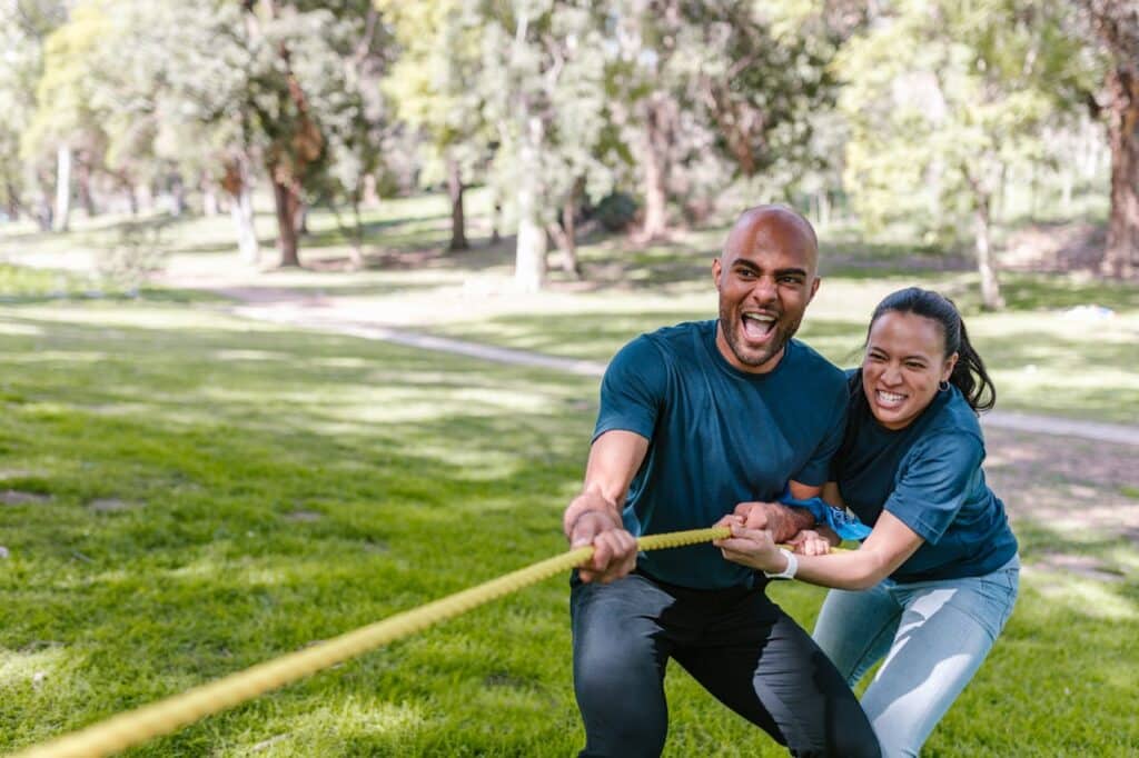 A man and woman in matching blue shirts, smiling and pulling a yellow rope in a tug-of-war game, with a backdrop of a lush park setting. Their expressions convey enjoyment and healthy competition in this outdoor team-building activity.