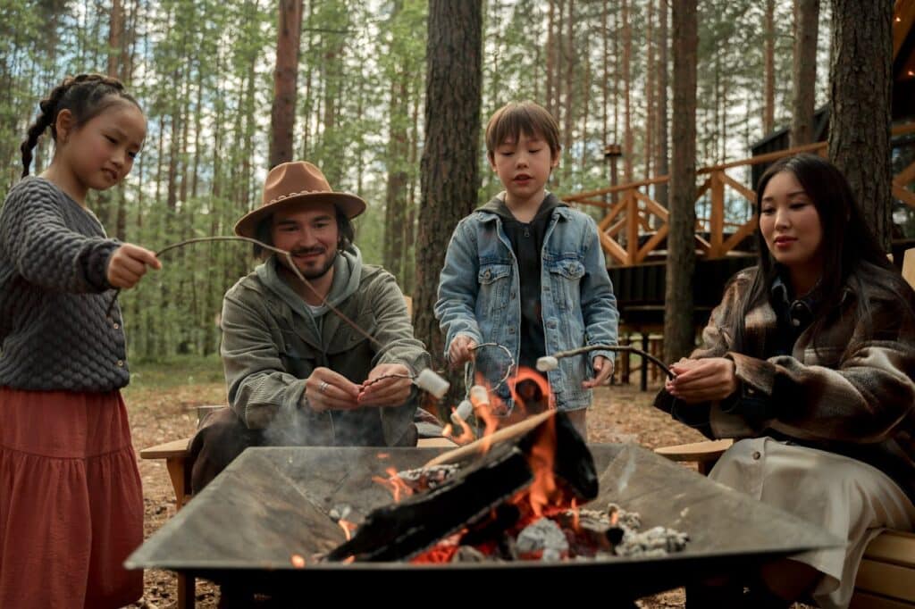 A family enjoying a campfire in a pine forest, with the father wearing a hat poking the fire, a daughter holding a skewer, and a son grilling marshmallows. The warm glow of the fire and the surrounding trees create a cozy and inviting outdoor setting.