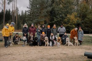A group of people and their dogs pose for a photo at a dog lovers' meet-up in a forest clearing. They are dressed in cozy autumn attire, with an array of colorful jackets, and the dogs vary in breed and size, looking up or at the camera attentively.