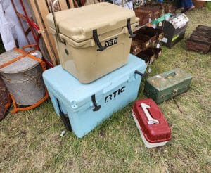 Two coolers, one beige Yeti and one light blue RTIC, are stacked on a grassy surface at an outdoor market, surrounded by various vintage metal containers and toolboxes