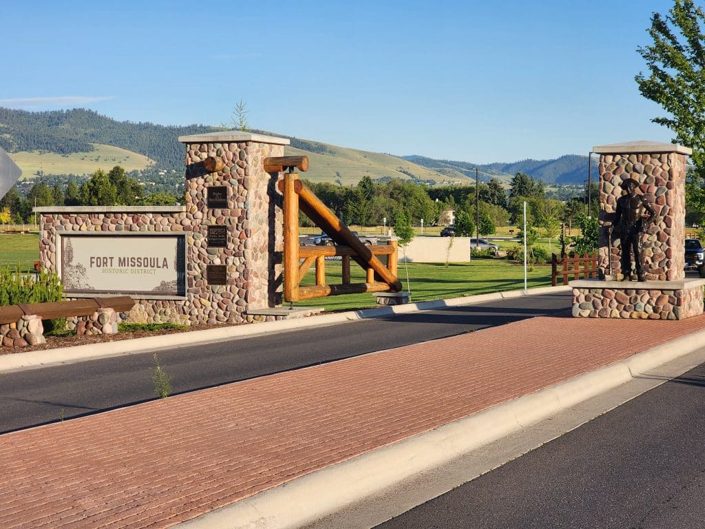 Entrance to Fort Missoula Historic District featuring a stone and timber gateway with a large sign and a bronze statue of a soldier, set against a backdrop of green fields and rolling hills under a clear blue sky.