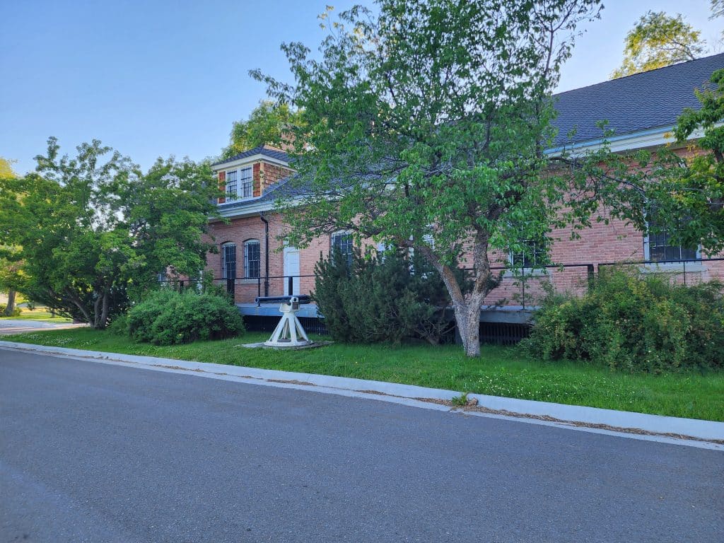 A historic brick building at Fort Missoula, partially obscured by lush green trees and bushes, with a well-maintained sidewalk and street in the foreground under a clear blue sky.