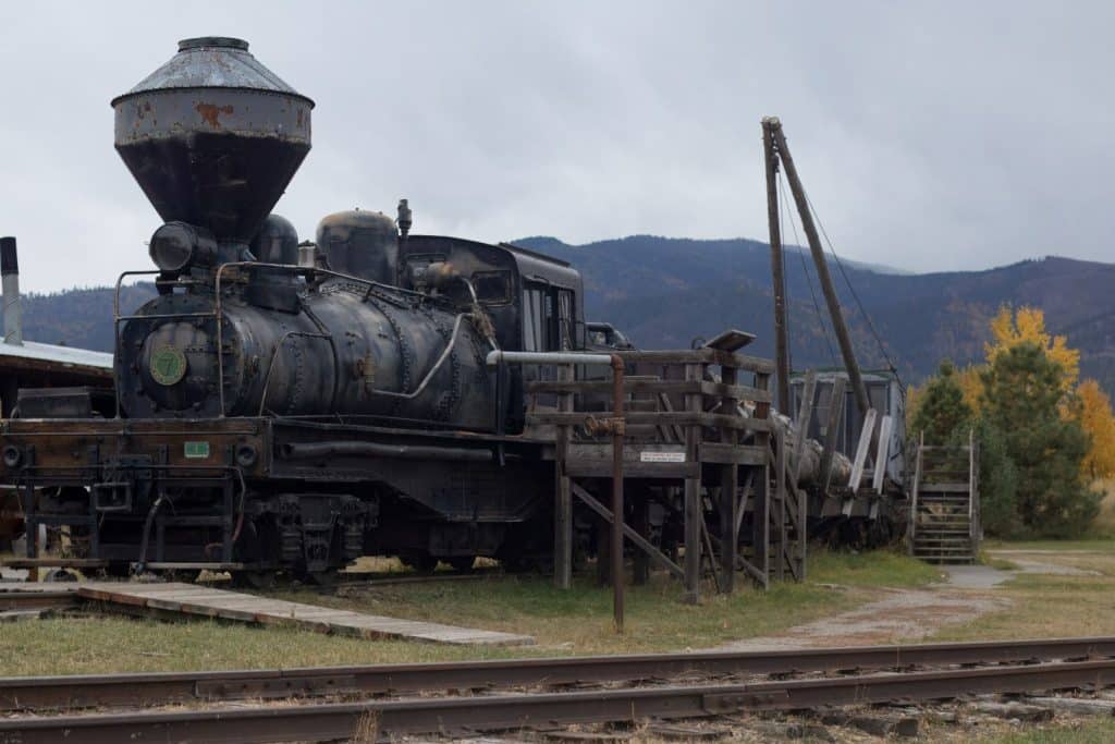 A vintage steam locomotive with the number 7 on its front stands idle on railway tracks, surrounded by wooden platforms and structures against a backdrop of misty mountains and autumnal trees.