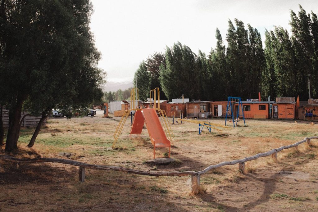 Rustic outdoor scene featuring an old playground with slides and swings, surrounded by a grassy area and tall trees. In the background, small wooden cabins can be seen under a cloudy sky.